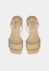 Sandales véganes beiges avec une large bride sur les orteils, une fine bride à la cheville avec une boucle, et une semelle plate. Texture lisse et design minimaliste.