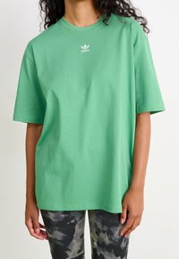 Osoba oblečená v volném zeleném tričku Adidas a černobílých legínách s tie-dye stojí proti jednobarevnému světlému pozadí.