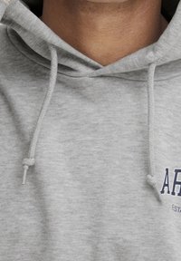Sudadera con capucha gris de tejido texturizado, con cordones y un logo azul marino en el pecho. Vista cercana de la zona del cuello.