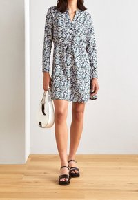 Vestido floral en azul, blanco y rosa con mangas largas, parte delantera abotonada y detalle de cintura atada. Combinado con un bolso blanco y sandalias negras.