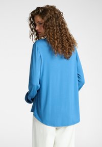 Camicia a maniche lunghe in tessuto blu brillante con una texture morbida, caratterizzata da un orlo curvato e maniche con polsini, indossata sopra dei pantaloni bianchi.