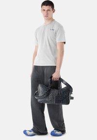 Joven de pie, vistiendo camiseta gris y pantalones, zapatos azules con textura, sosteniendo un gran bolso oscuro con estampado por las asas.