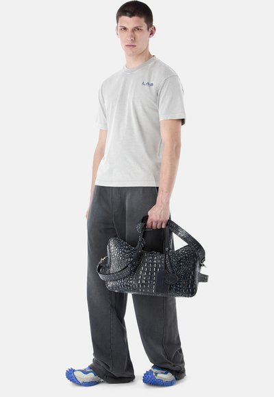 Jeune homme debout, portant un t-shirt gris et un pantalon, des chaussures bleues texturées, tenant un grand sac à main foncé à motifs par ses poignées.