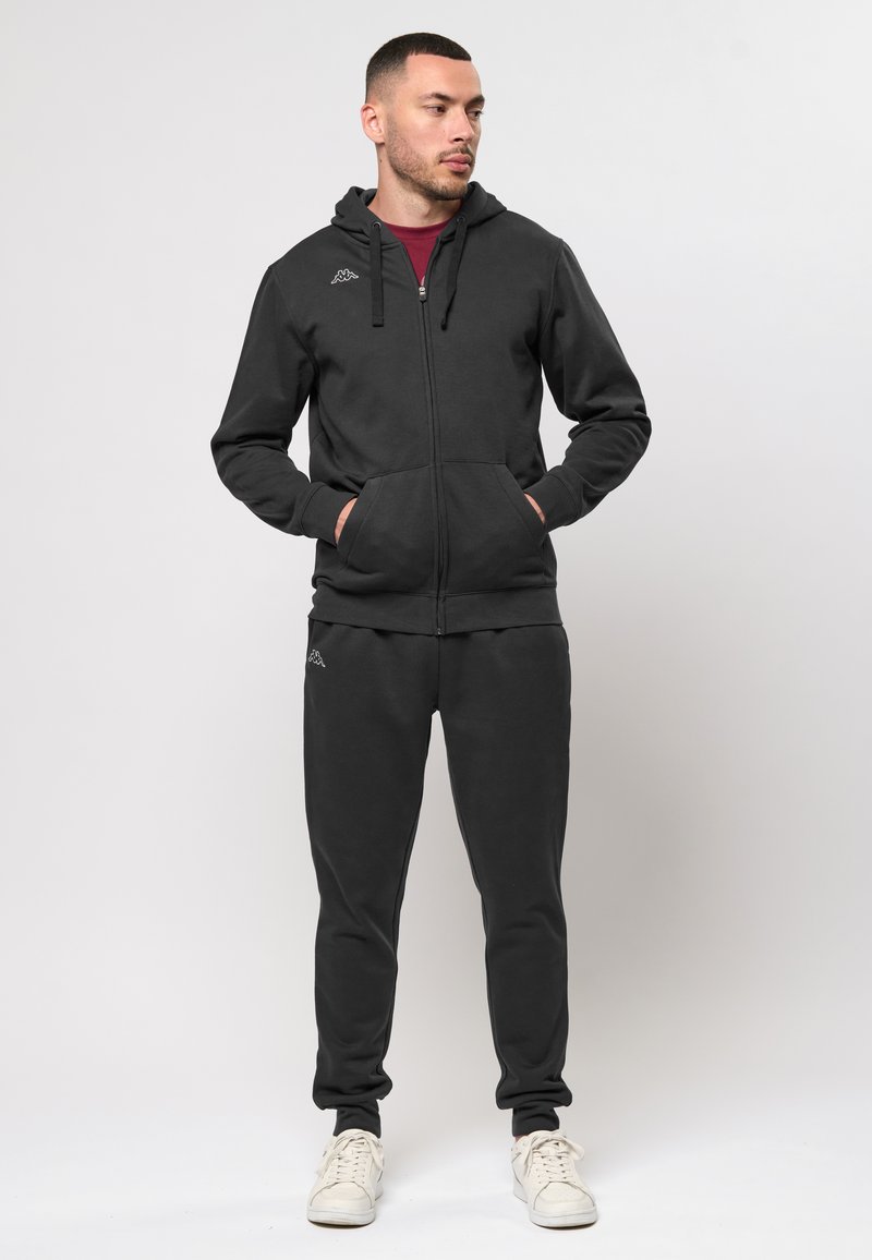 Fekete, kapucnis, cipzáros kapucnis dzseki, elülső zsebekkel és szürke részletekkel, fekete, puha, text�úrált anyagú joggerváltozattal párosítva.