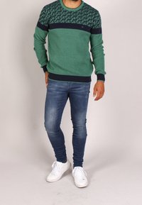 Zielony sweter z wzorzystą górną częścią, jednolita granatowa wstążka pośrodku, dzianinowa struktura, w połączeniu z wąskimi dżinsami i białymi trampkami.