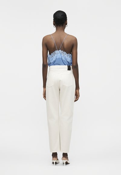 Ženska s temno kožo in lasmi v čopku, oblečena v modro majico z čipkastim robom, bele hlače z visokim pasom in bele sandale z visokimi petami, obrnjena stran.