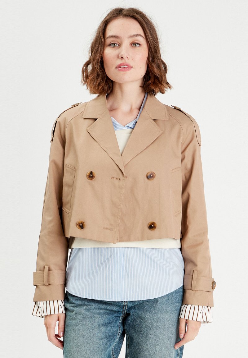 Veste beige cropped à double boutonnage avec des boutons marron, épaulettes et manchons rayés contrastants sur une chemise bleu clair.
