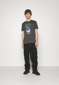 T-shirt en coton gris foncé avec un graphique NASA en vert fluo et bleu. Associé à un pantalon large noir et des chaussures noires.