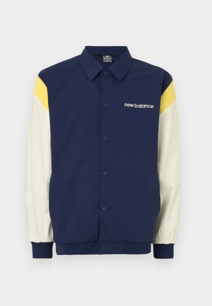 Chaqueta azul marino con acentos en amarillo y blanco, cierre de botones, mangas largas y un logo en el pecho. Cuenta con bolsillos laterales.