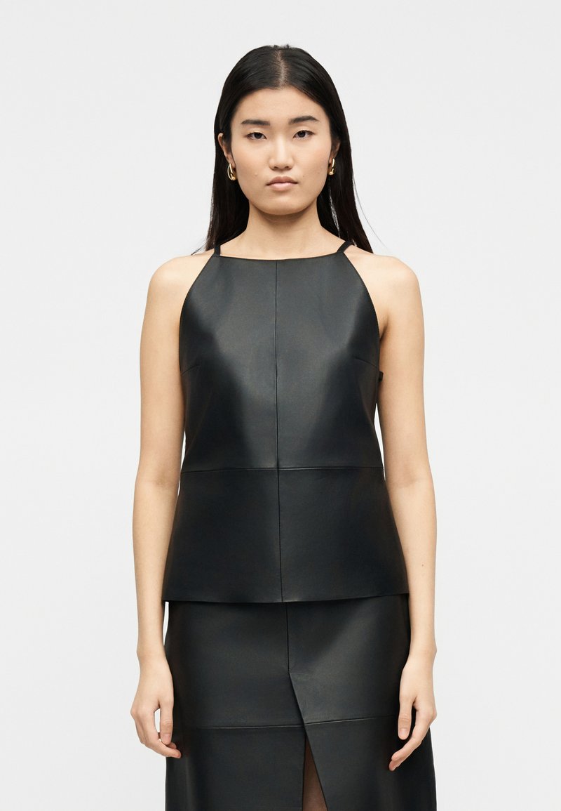 Γυναίκα με ίσια μαύρα μαλλιά που φορεί χρυσούς κρίκους στα αυτιά, κορυφή από μαύρο δερμάτινο χωρίς μανίκια και ταιριαστό φούστα με σχίσιμο μπροστά, στέκεται μπροστά σε λευκό φόντο.