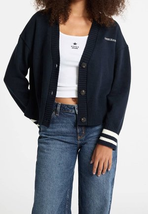 Femme portant un cardigan bleu marine boutonné avec des rayures blanches aux poignets, par-dessus un débardeur blanc court et un jean bleu.