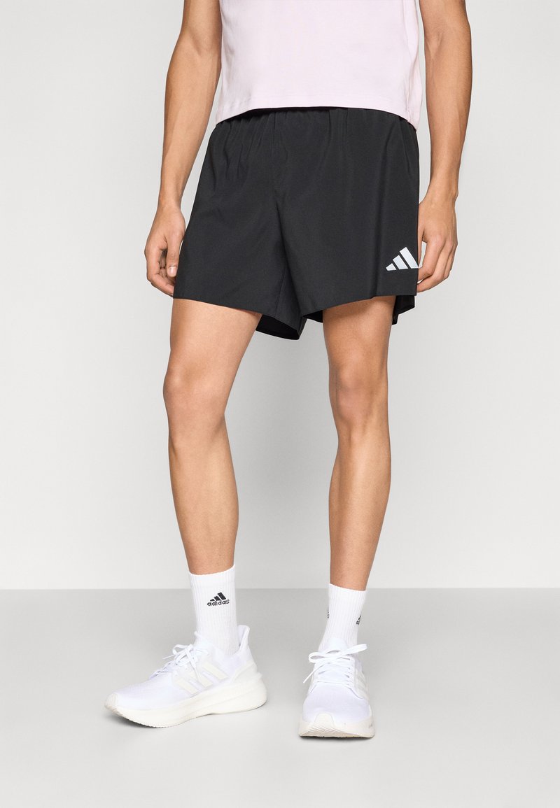 Fekete atlétikai rövidnadrág rugalmas derékpánttal és három fehér csík díszítéssel a bal lábszáron, fehér sportcipővel és zoknival párosítva.