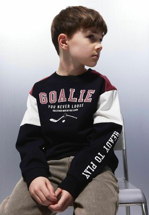 Črna in mornarsko modra jakna s burgundskimi in belimi akcenti, ki prikazuje besedilo "GOALIE" in grafiko hokejista. Mehka, bombažna tkanina z rebrastimi manšetami.
