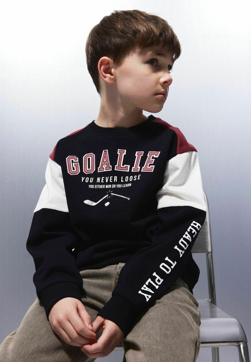 Juoda ir tamsiai mėlyna megzta palaidinė su bordo ir baltais akcentais, su užrašu "GOALIE" ir ledo ritulio grafika. Minkšta, medvilninė medžiaga su gumuotais rankogaliais.