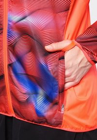 Main dans la poche d'une veste orange avec un motif ondulé noir, bleu et rose et une boucle pour le pouce sur la manche près de la fermeture éclair.