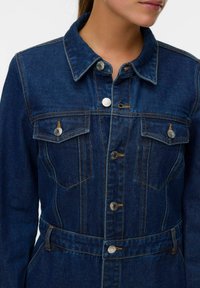 Jeans jakna temno modre barve z gumbom spredaj, dvema žepoma na prsih in pasom z zaponko. Ima kontrastno šivanje in kovinske dodatke.