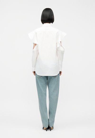 Femeie cu păr scurt și negru, purtând o bluză albă cu mâneci lungi și umeri decupați și blugi albaștri deschiși cu croială dreaptă, stând cu spatele către cameră.