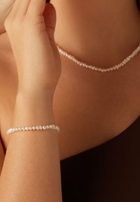 Parelsieraad en armband set met kleine, ronde witte parels met een gladde textuur, gedragen om de nek en pols tegen een donkere achtergrond.