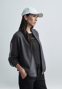 Szara kurtka typu bomber rozpinana na zamek z białymi paskami, w połączeniu z czarnym t-shirtem i czarnymi spodniami. Biała czapka baseballowa dopełnia stylizację.