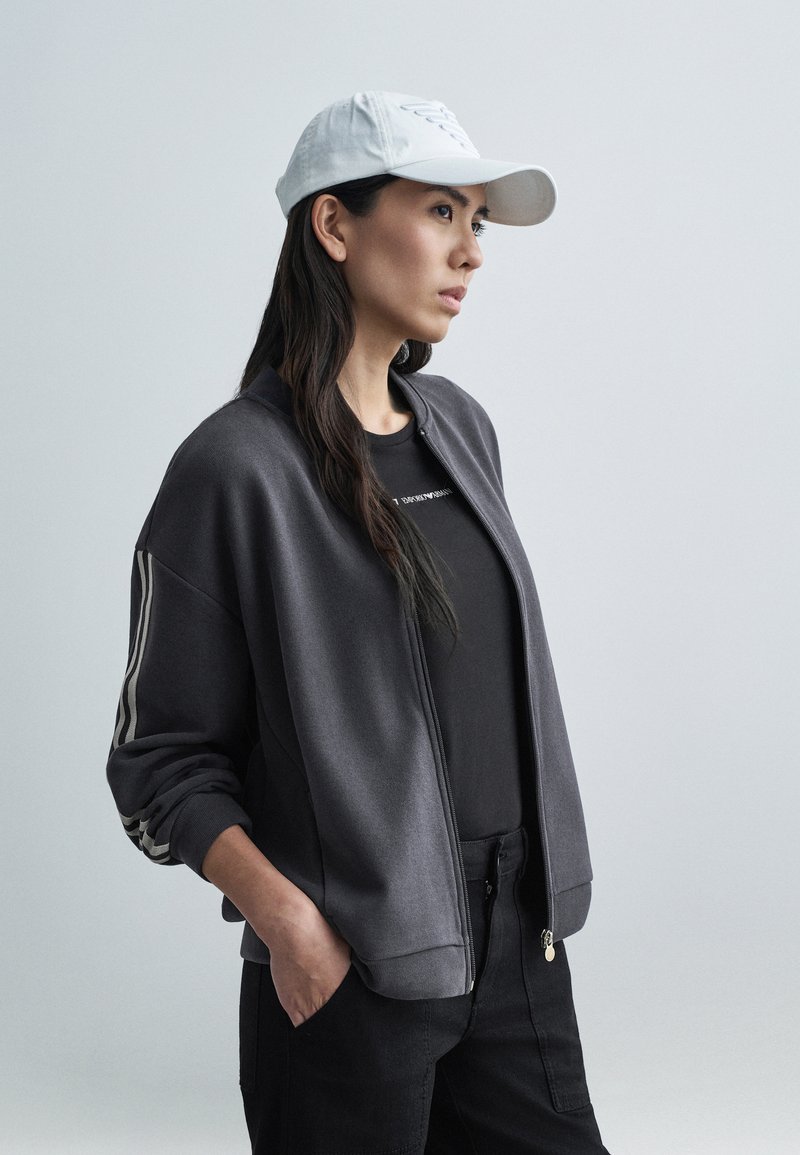 Szara kurtka typu bomber rozpinana na zamek z białymi paskami, w połączeniu z czarnym t-shirtem i czarnymi spodniami. Biała czapka baseballowa dopełnia stylizację.