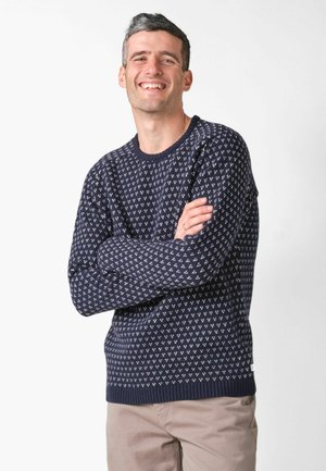 Usmievavý muž s krátkymi tmavými vlasmi, oblečený v námornícky modrom vzorovanom svetri a béžových nohaviciach, stojaci so založenými rukami na pozadí bielej steny.