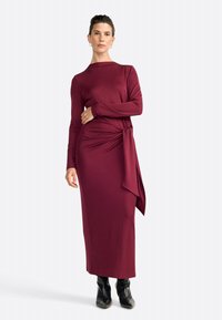 Bordeauxfarbenes Langarmkleid mit figurbetonter Silhouette, seitlichem Knotendetail und weichem Material, kombiniert mit schwarzen Knöchelstiefeln.