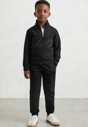 Melnā sporta treniņtērps ar aizdari pie kakla, kura priekšējā daļā ir teksturēta melna paneļa un pieguļoši bikses. Apavi ir balti ar bežām akcentiem.