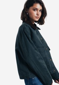 Mörkgrön jacka i ett hållbart tyg med en brun kordflöjtskrage, tryckknappar och sidofickor; avslappnad, oversized passform.