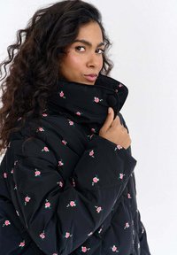 Czarna pikowana kurtka typu puffer z haftem kwiatowym, wysokim kołnierzem i detalem zapinanym na guziki. Pikowany materiał z drobnymi wzorami czerwonych i niebieskich róż.