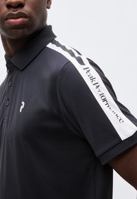Homme portant un polo noir avec une bande blanche à l'épaule comportant le texte et le logo « Peak Performance » sur la poitrine.