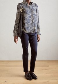 Blusa navy con pois beige, design con bottoni e maniche lunghe; abbinata a pantaloni navy su misura e mocassini neri. Cinghia della borsa nera visibile.