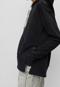 Osoba oblečená v černé mikině s bílými šňůrkami a bočním zipem, s rukama v kapsách, doplněná o světle šedé džíny.