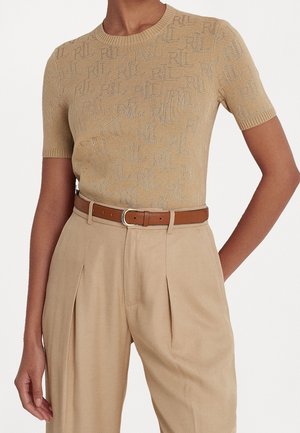 Femme portant un haut en maille beige à manches courtes avec un motif subtil, rentré dans un pantalon beige taille haute avec plis et une ceinture en cuir marron.