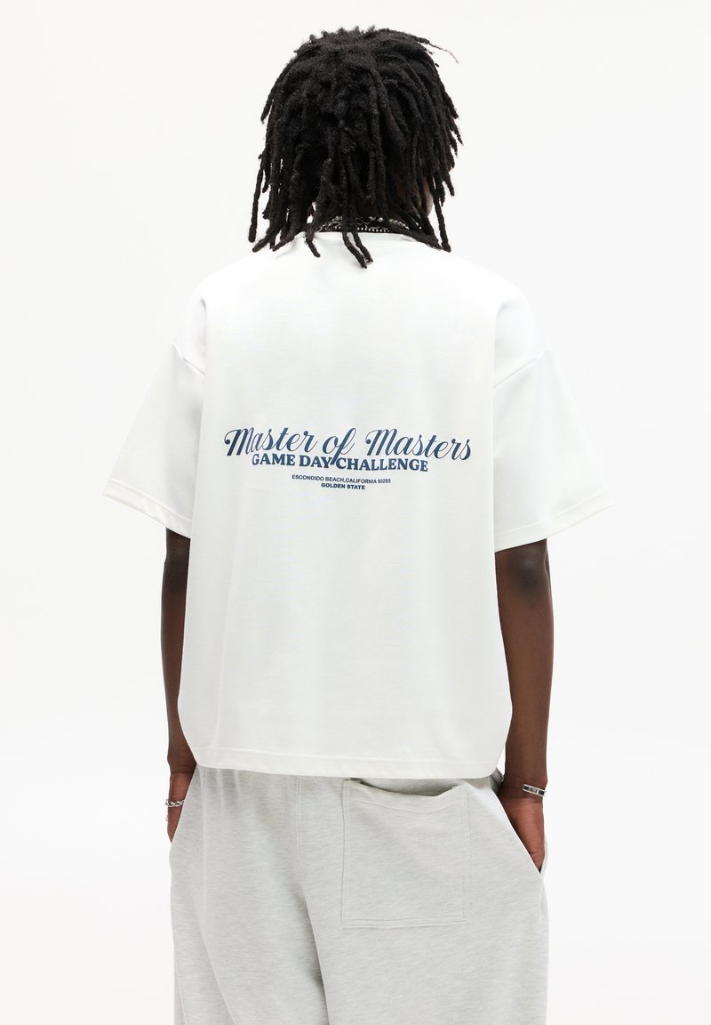 Hvid oversized t-shirt med "Master of Masters Game Day Challenge" trykt i blåt på ryggen. Blødt bomuldsstof og afslappet pasform.