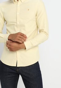 Helles gelbes Langarmhemd mit Button-Down-Kragen, mit einem kleinen gestickten Logo auf der Brust, kombiniert mit dunkelblauen Jeans.