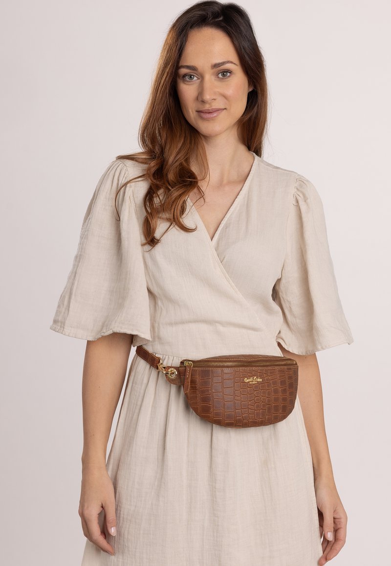 Marsupio in pelle stampata coccodrillo marrone con cerniera dorata, indossato in vita sopra un vestito in lino beige con maniche corte e a sbuffo.