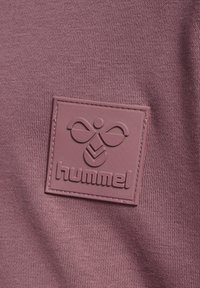 Zbliżenie fioletowej tkaniny z naszytym gumowym naszywką przedstawiającą stylizowane logo pszczoły i nazwę marki "hummel".