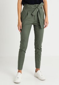 Olivgrön högmidjad byxor med bälte i midjan, framfällt och avsmalnande ben, i kombination med vita sneakers.