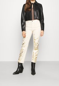Chaqueta recortada de cuero negra, top marrón, pantalones crema con estampado abstracto dorado y botas de tobillo negras con punta.