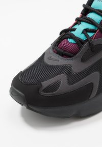 Lähedalt vaadates must ja hall spordijalats, millel on maroonivärvine võrk, tealist keelt ja mustad sooned valgel taustal.