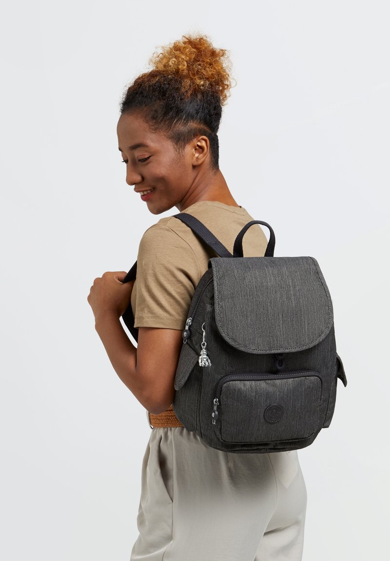 Sac à dos en tissu gris avec rabat repliable, poche avant et charm décoratif. Dispose de bretelles ajustables et d'un design épuré.