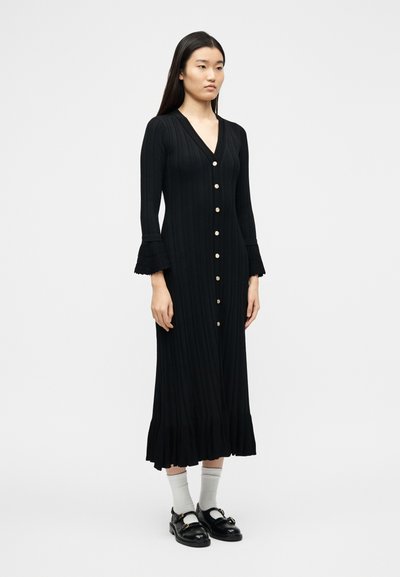 Γυναίκα που φοράει μαύρο, μακρυμάνικο midi φόρεμα με κουμπιά και φαρδ ιές μανσέτες, συνδυασμένο με λευκές κάλτσες και μαύρα loafers, στέκεται μπροστά σε λευκό φόντο.