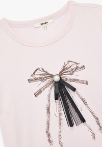 Camiseta de manga corta rosa claro con un gráfico de lazo en blanco y negro. Presenta un acento de perla y detalles de cinta negra. Hecha de algodón suave.