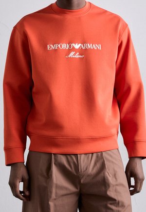 Muž nosící oranžovou mikinu s dlouhým rukávem s logem "Emporio Armani Milano", v kombinaci s hnědými kalhotami, stojí před jednobarevným pozadím.