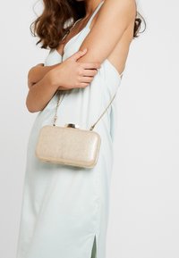 Bolso de mano metálico dorado con forma rectangular, superficie texturizada y una cadena como asa. El cierre es un broche redondeado en la parte superior.