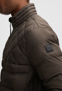 Giacca imbottita verde oliva scuro, con collo alto, motivo trapuntato a losanghe e una toppa con logo sulla spalla sinistra.