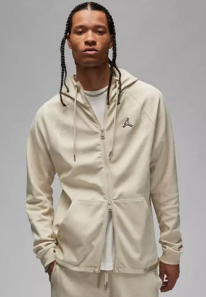 Uomo con capelli intrecciati indossa una felpa beige con zip e logo Air Jordan e pantaloni abbinati, in piedi davanti a uno sfondo grigio uniforme.