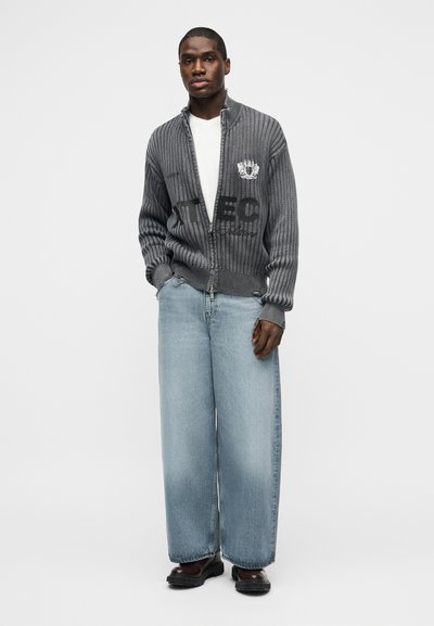 Άνδρας που φοράει γκρι πλεκτό πουλόβερ με φερμουάρ και λογότυπο, λευκό μπλουζάκι, φαρδιά ανοιχτό μπλε τζιν και σκούρα καφέ παπούτσια, στέκεται με τα χέρια στις τσέπες.