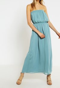 Femme portant une robe maxi sans bretelles, bleu clair, associée à des talons nus à bout ouvert, se tenant devant un fond blanc uni.