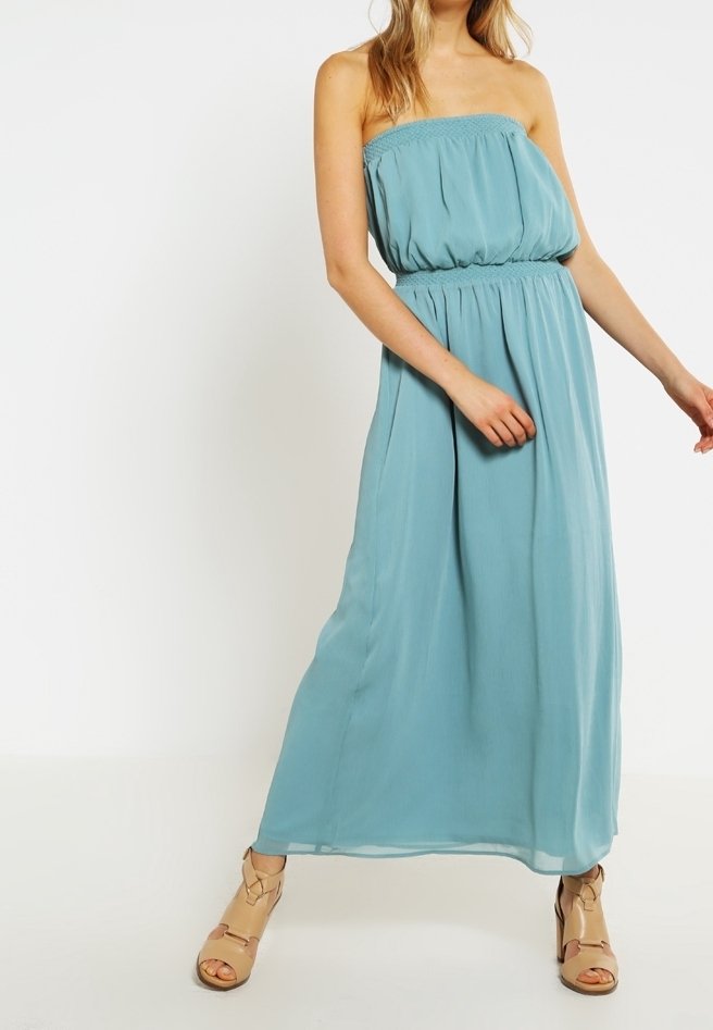 Femme portant une robe maxi sans bretelles, bleu clair, associée à des talons nus à bout ouvert, se tenant devant un fond blanc uni.